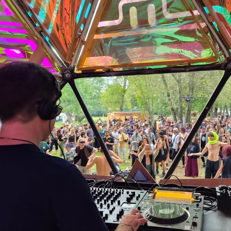 Neurodriver behind the decks, looking out through a geometric stage canopy at a festival crowd dancing in woodland.
