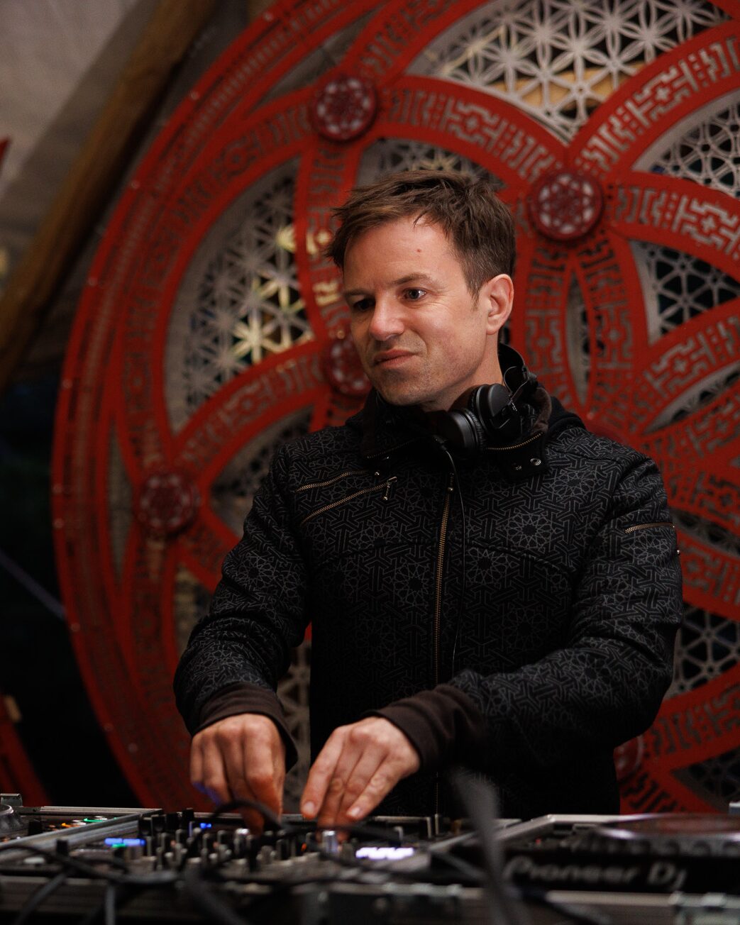 Neurodriver playing a DJ set at Magikana, headphones round his neck and hands on the mixer, framed by a large carved red mandala backdrop.